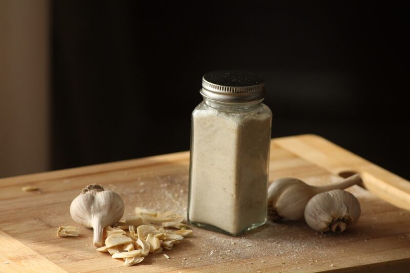 on a wooden board is spice jar full of garlic powder, cloves of garlic, and dried garlic chips