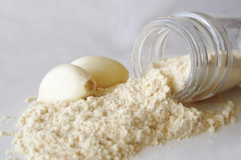 on a white surface is toppled over jar of garlic powder with a couple of peeled garlic cloves