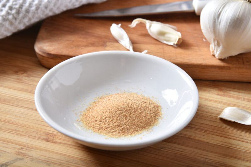 on a wooden surface is a white ceramic dish with a little mound of garlic powder, with a wooden chopping board behind it with a couple of cloves and a head of garlic