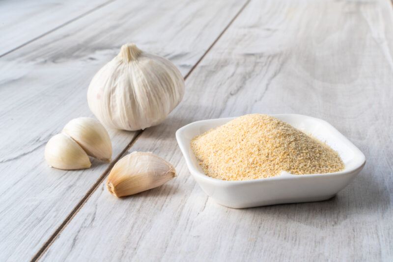 on a white wooden surface is a white square dish with garlic powder, beside is a head and 3 cloves of fresh garlic