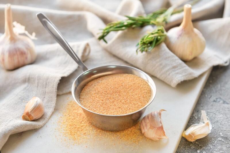 on a white board is a metal scoop full of garlic powder, around it are loose garlic powder with beige table napkin with garlic cloves on it and fresh herbs