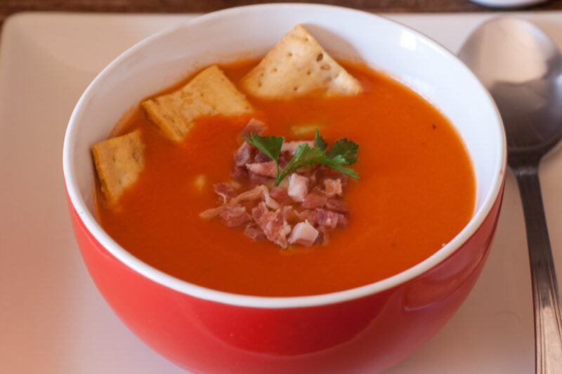 closeup image of a red and white bowl with gazpacho, a cold soup, with croutons, bacon bits, and sprig of herb for toppins