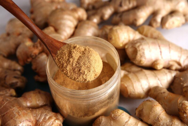 a wooden spoon full of ginger powder over a plastic jar with ginger powder as well, around it are fresh ginger rhizomes