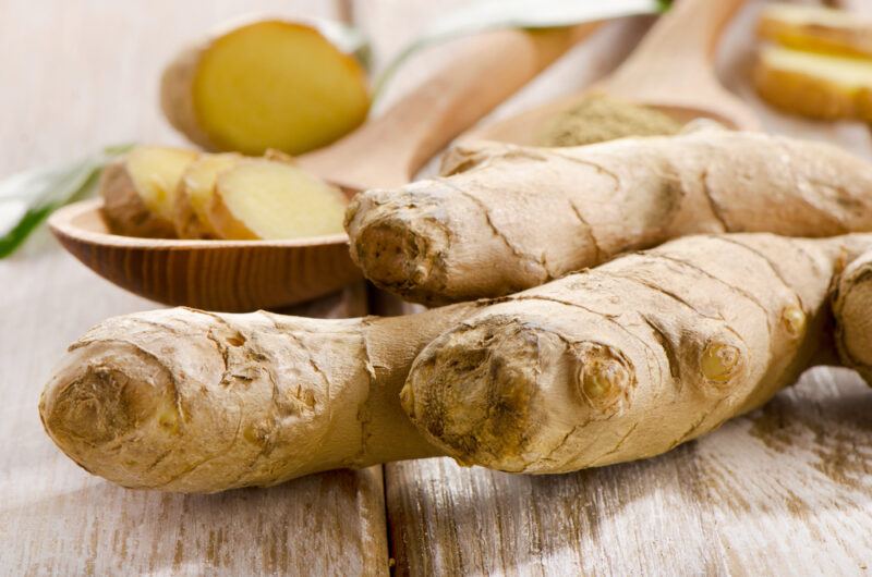 on a rustic wooden surface is a fresh ginger rhizome, at the back are two wooden spoons with sliced ginger and powdered ginger