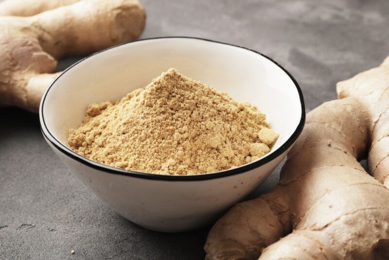 on a concrete surface is a white bowl with blue rim full of ginger powder, beside it are fresh ginger rhizomes