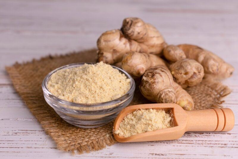 on a wooden surface is a burlap with a clear glass bowl of ginger powder with a wooden scoop beside it with ginger powder as well, at the back are fresh ginger rhizomes