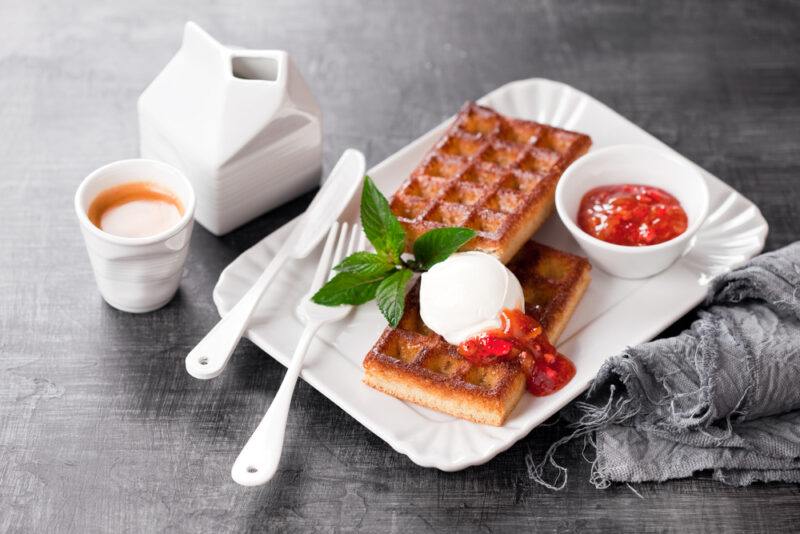 on a black concrete surface is a rectangular white dish with beautifully plated grain-free waffles with a dollop of cream, strawberries, and sprigs of herbs on top, beside is a silver knife and fork, a gray table napkin on one side, a cup of tea and carton of milk on the other side