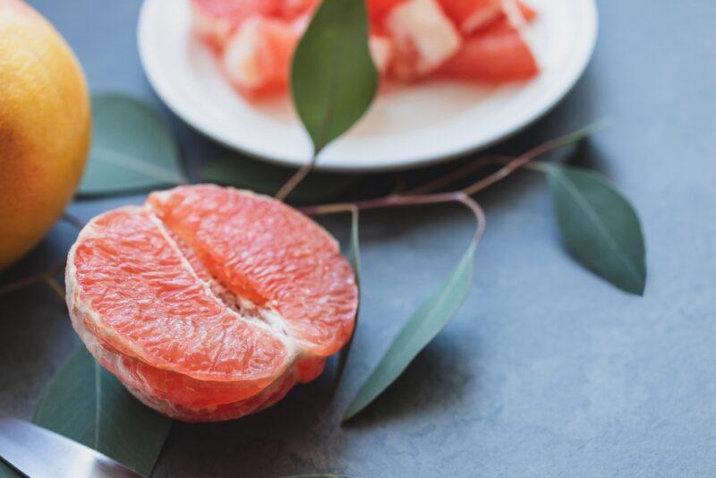 on a black concrete surface is a halve and peeled grapefruit with a small white dish behind it with grapefruit bits and a thin stem with a few leaves