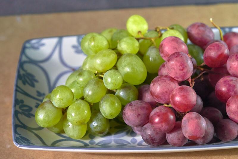 on a wooden surface is a rectangular white dish with blue designs with a couple of bunches of green and red grapes