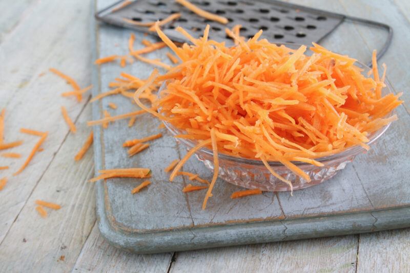 on a rustic looking wooden surface is a faded blue wooden board with a glass of bowl with grated carrots, at the back is a metal grater, around it are loose grated carrots