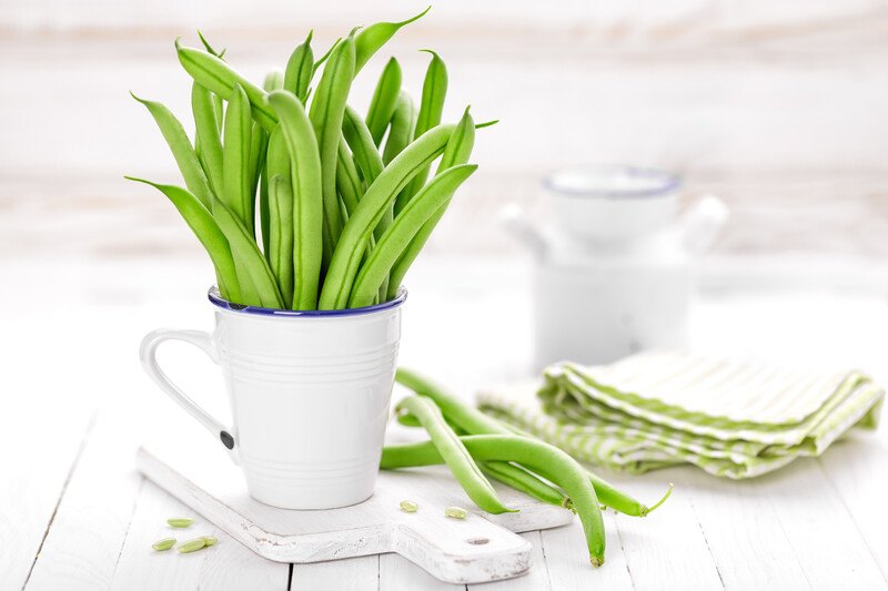 on a white wooden surface is a white chopping board with a white mug full of green beans with green beans at the back with green and white kitchen towel