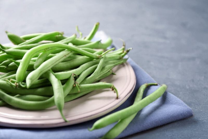 on a grey concrete surface is a dish full of green beans resting on a blue table napkin