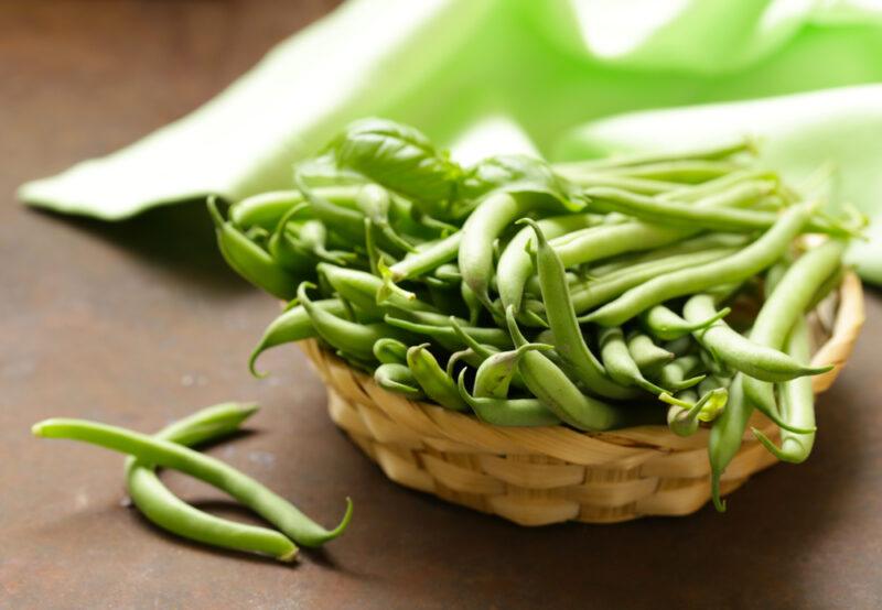 on a brown surface is a weaved shallow basket full of green beans, with a couple of green beans beside it and a green table napkin at the back