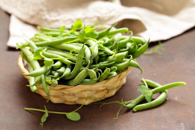 on a concrete surface is a basket full of green beans with loose green beans beside it and a white kitchen towel at the back