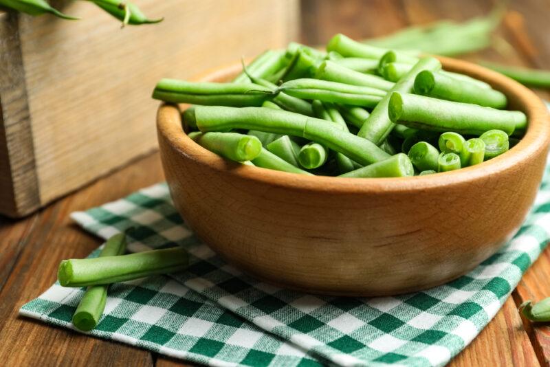 on a wooden surface is a wooden bowl full of cut green beans resting on top of a white and green checkered table napkin, with a loose green bean beside it, at the back is a brown wooden box with green beans as well