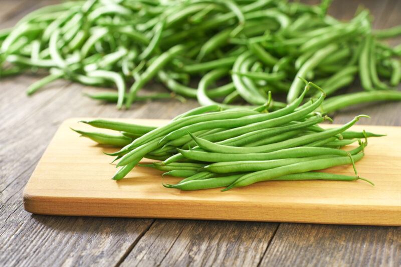 on a wooden surface is a wooden chopping board with green beans on it, at the back is a pile of fresh green beans