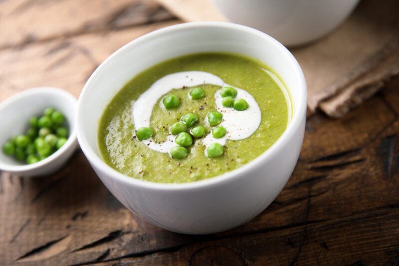 on a rustic looking wooden surface is a white bowl of green pea soup with a few green peas on top, beside it is small dish of green peas as well