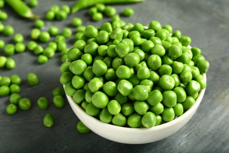 on a grey concrete surface is a white bowl full of green peas with loose green peas around it