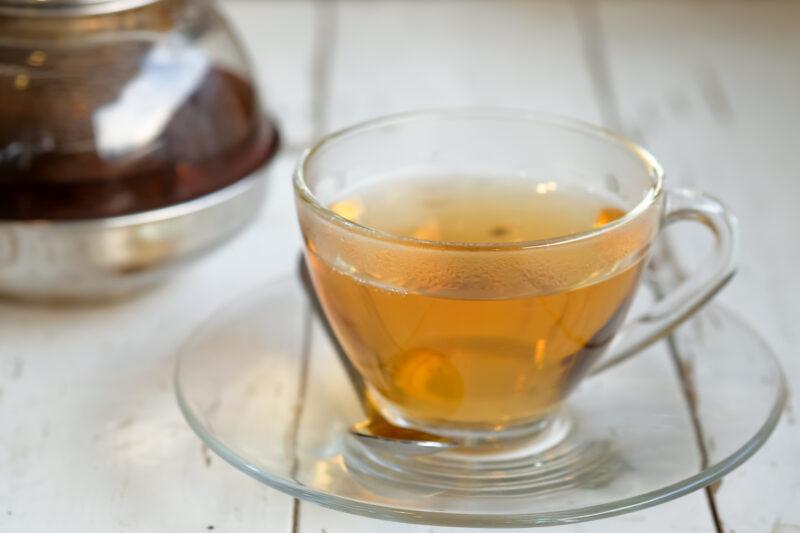 on a white wooden surface is a clear glass cup and saucer with green tea, behind it is a clear glass teapot with green tea as well