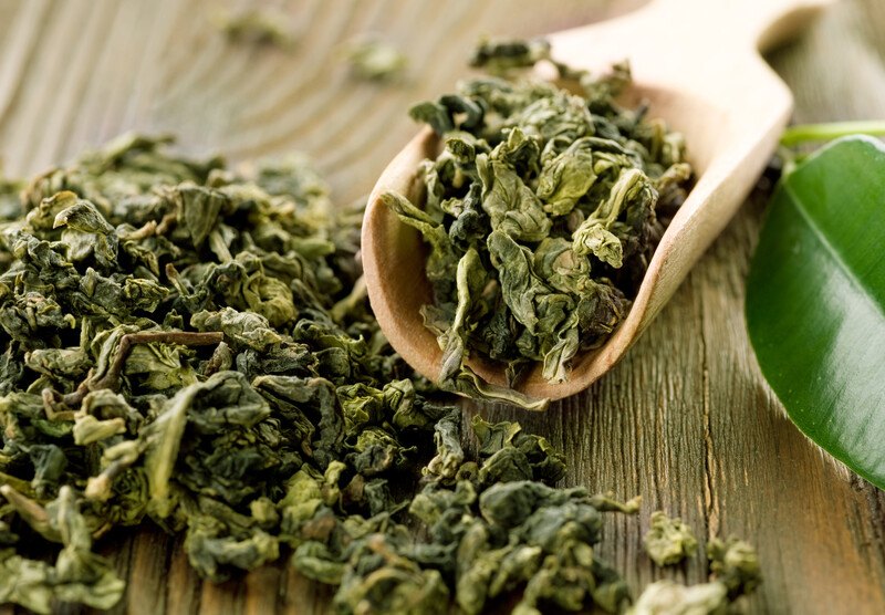 on a wooden surface is a closeup image of organic tea leaves with a wooden scoop full of green tea leaves