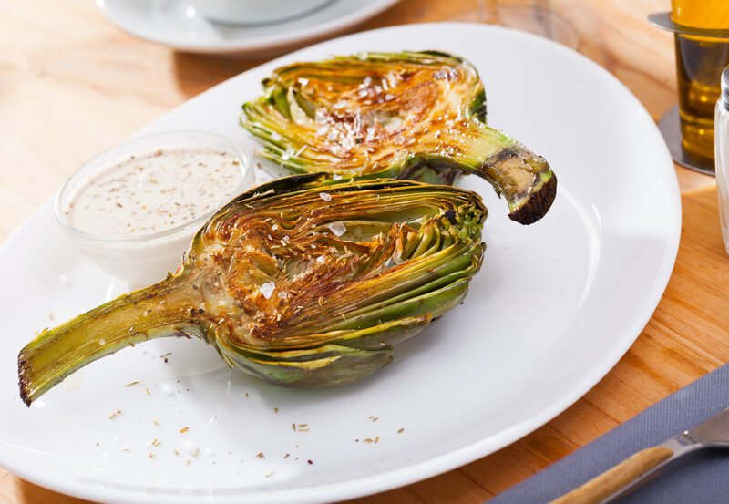 closeup image of a grilled halves artichoke on a white plate with a small glass bowl with white dipping sauce