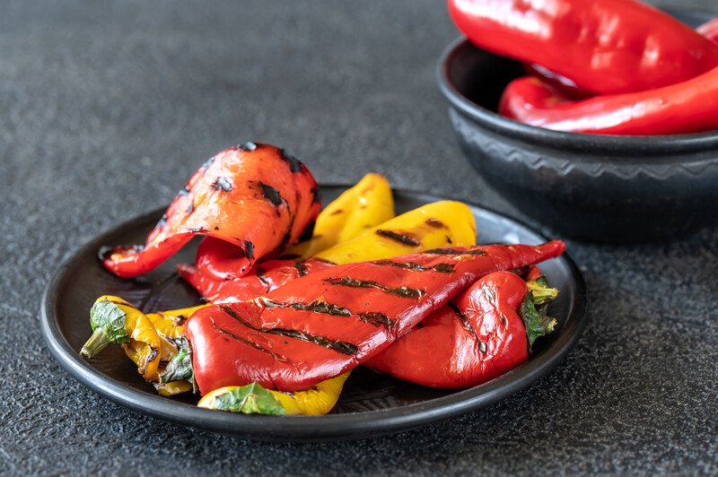 on a black surface is a black dish with grilled red and yellow bell peppers, with black dish full of fresh red bell peppers at the back