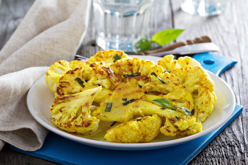 grilled yellow cauliflower on a white dish on a blue wooden chopping board with white table napkin and glass of water