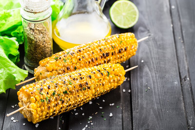 on a black wooden surface is a couple of grilled corn on sticks, sprinkled with rock salt, with leafy greens, small jar of dried herbs, olive oil, and a sliced lime at the back