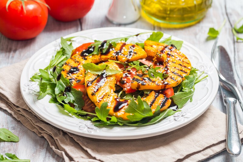 a beautifully plated grilled squash with herbs, greens, and balsamic dressing, plate is resting on a brown table napkin with knife and fork beside it and a couple of tomatoes and olive oil at the back