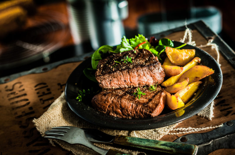 grilled steak served with potato wedges | Food For Net two pieces of steak cooked medium rare, sered on a black plate with a side of roasted potato wedges.