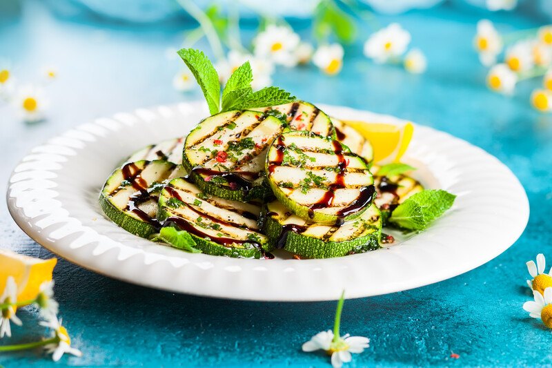 on a blue surface is a beautifully plated grilled zucchini with a sprig of herb and white little flowers around the white plate