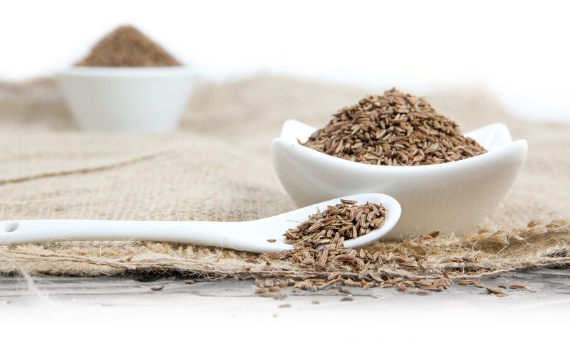 a white spoon and bowl full of cumin seeds with a bowl of ground cumin at the back, all resting on a burlap, on top of a white surface