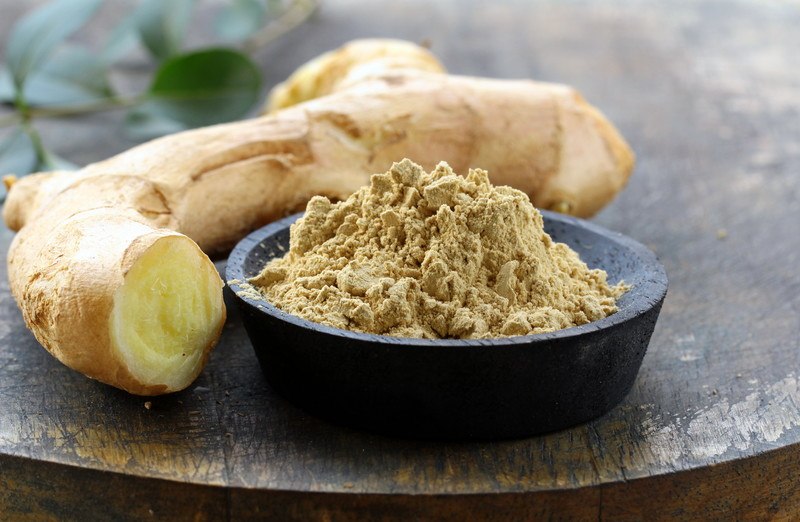 ground ginger in a black bowl on a wooden surface with fresh ginger beside it.