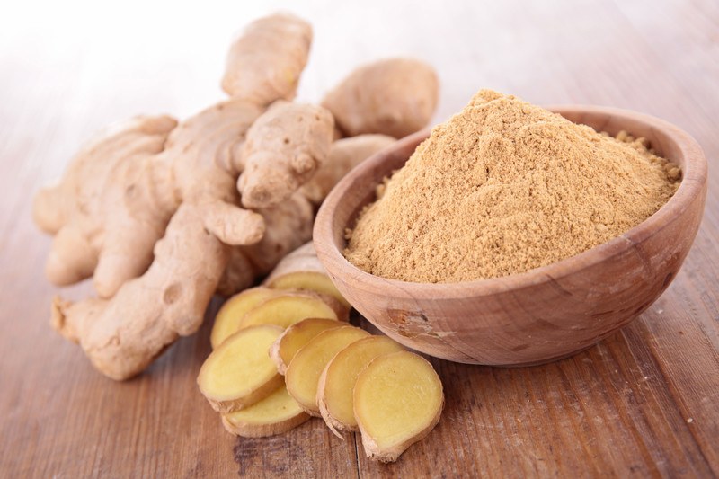 a wooden bowl full of ground ginger resting on a wooden surface with fresh ginger and sliced fresh ginger beside it