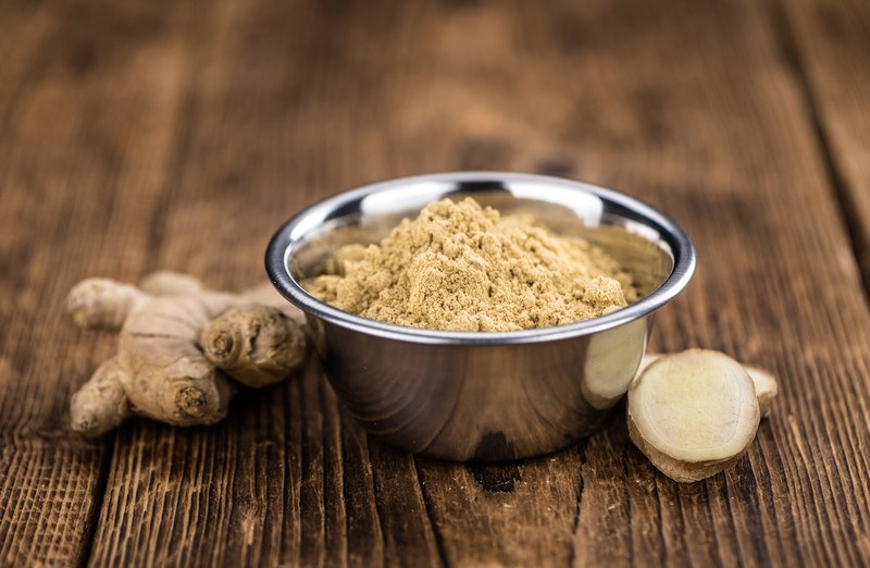 on an aged wooden surface is a stainless steel bowl full of ground ginger, with fresh ginger beside it