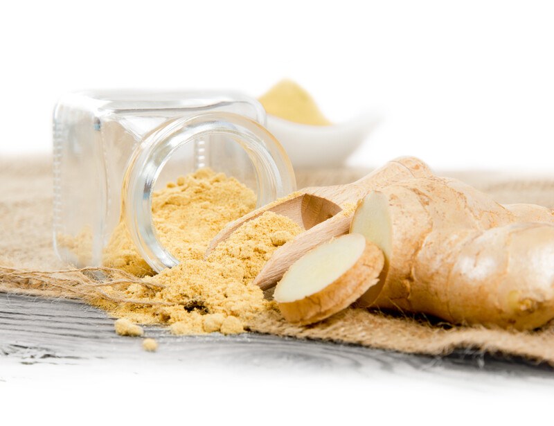 a little glass jar toppled over a burlap sack with ground ginger spilled from it, with wooden scoop also with ground ginger, fresh ginger root, and white bowl with ground ginger at the back