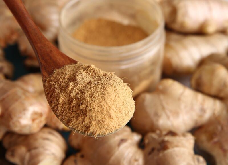 a closeup image of fresh ginger rhizomes with a plastic jar full of ground ginger, over them is a wooden spoon full of ground ginger as well