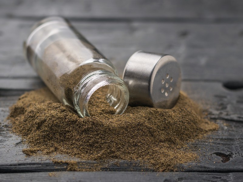 a small glass spice bottle with ground pepper spilled over on a black wooden surface 