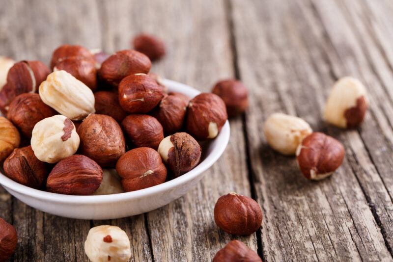 on a rustic wooden surface is a white dish full of hazelnuts, around it are loose hazelnuts