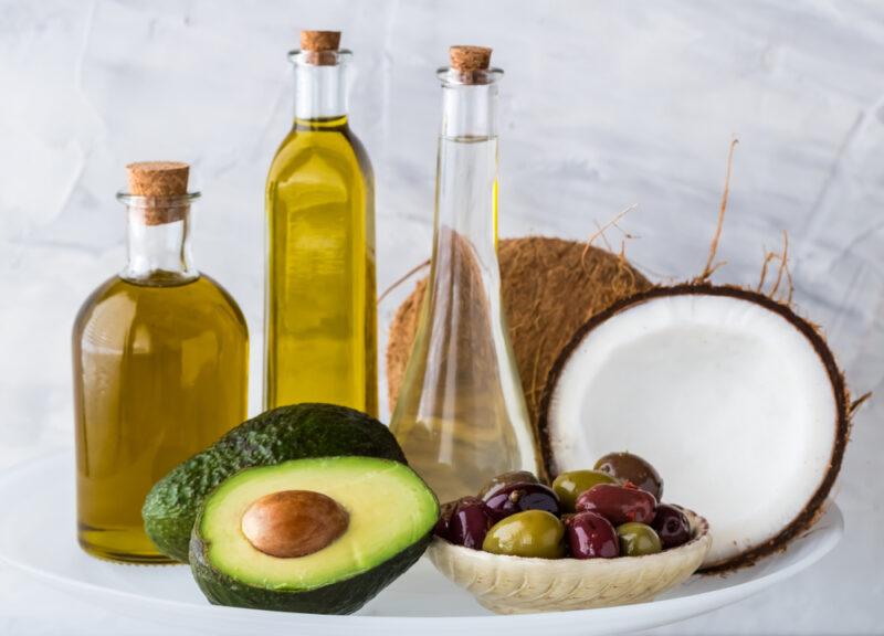 on a white big platter are different bottles with different healthy oils with halves coconut at the back and a small dish of assorted olives and avocados at the foreground