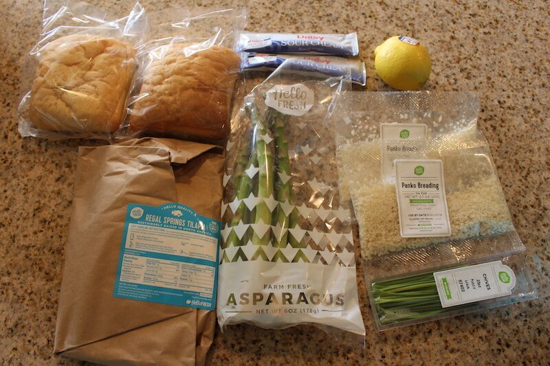 fresh ingredients sitting on counter including ciabatta rolls, lemon, sour cream, panko breading tilapia, and asparagus