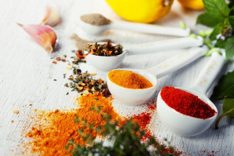 a closeup image of ceramic measuring spoons with spices and herbs lined up on a white surface, with loose spices and herbs around it