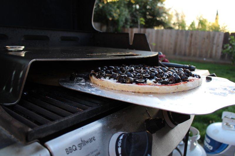 putting homemade olive pizza into gas grill with kettlepizza and pizza stone