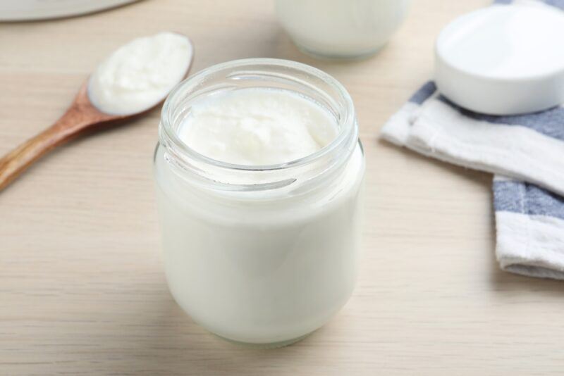 on a wooden surface is a jar of home made yogurt, at the back is a wooden spoon with yogurt, a partial glass with yogurt as well, and white and blue table napkin with a white lid for the jar