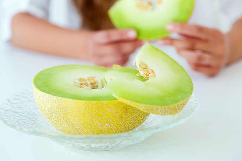 on clear glass dish is a halve honeydew melon, at the back is a partial image of hands holding a slice of honeydew melon