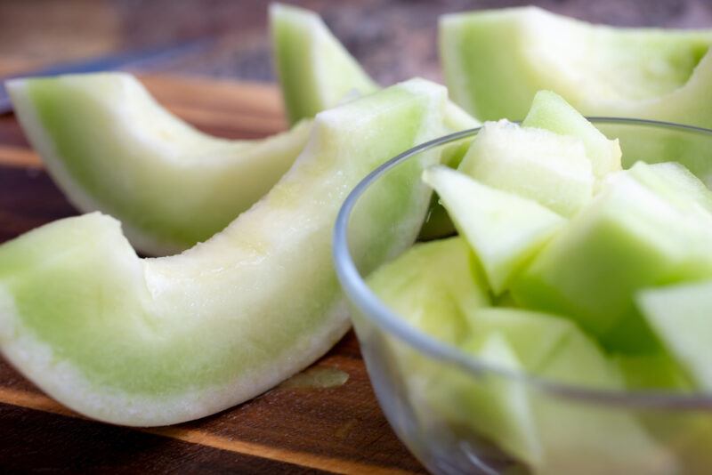 on a wooden surface is a closeup image of sliced honeydew melon with a clear glass bowl with honeydew melon bits