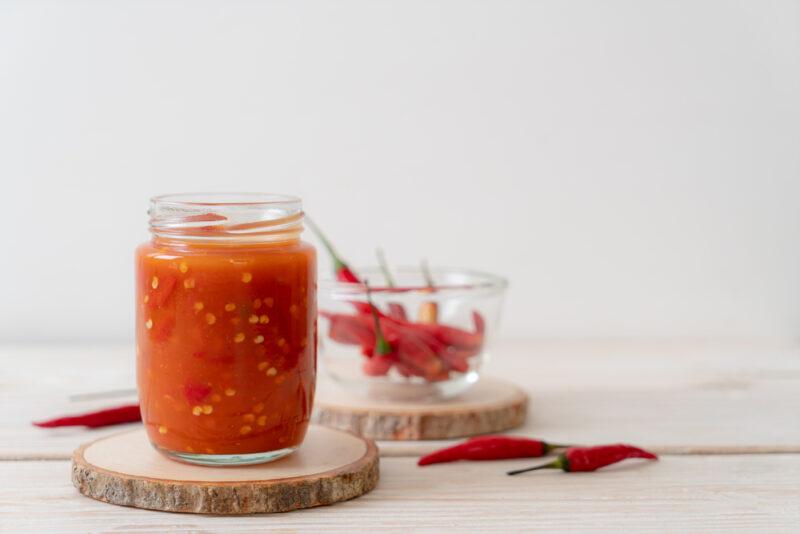 a jar of hot sauce resting on a round block of wood, beside it is a few fresh chili peppers, at the back is a small clear bowl with fresh chili peppers as well