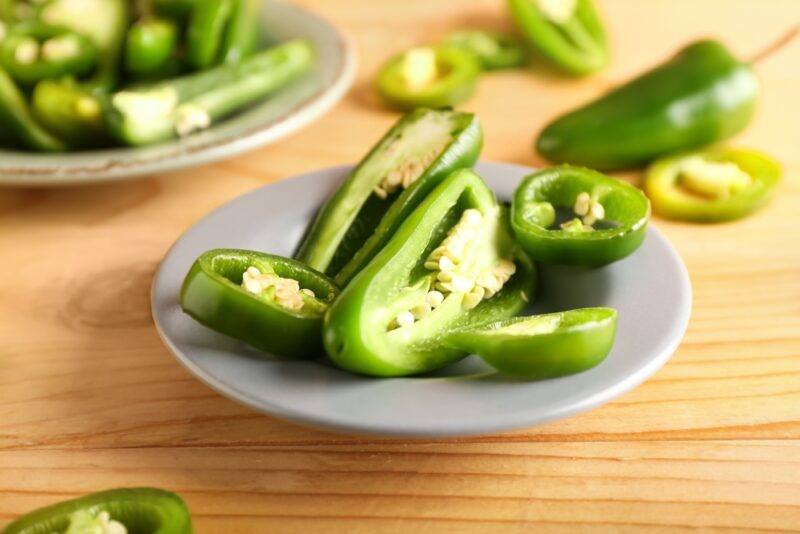 on a wooden surface is a small gray glass dish with sliced jalapeno, around it are loose jalapeno peppers, at the back is a ceramic dish also with sliced jalapeno peppers