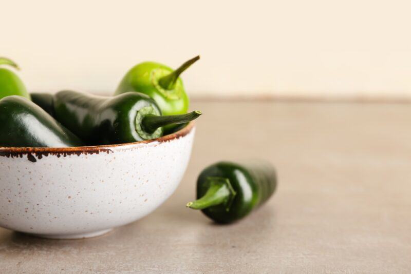 on a beige colored surface is a white ceramic bowl with brown specs and brown rim , full of jalapenos, beside it is one loose jalapeno pepper