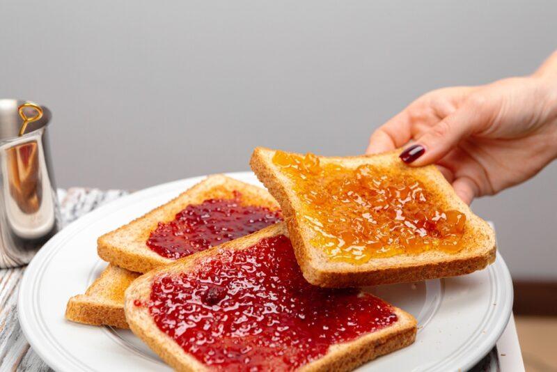 pile of toasts on a white plate with different kinds of jams, a partial image of a hand can be seen taking one of the toast from the plate